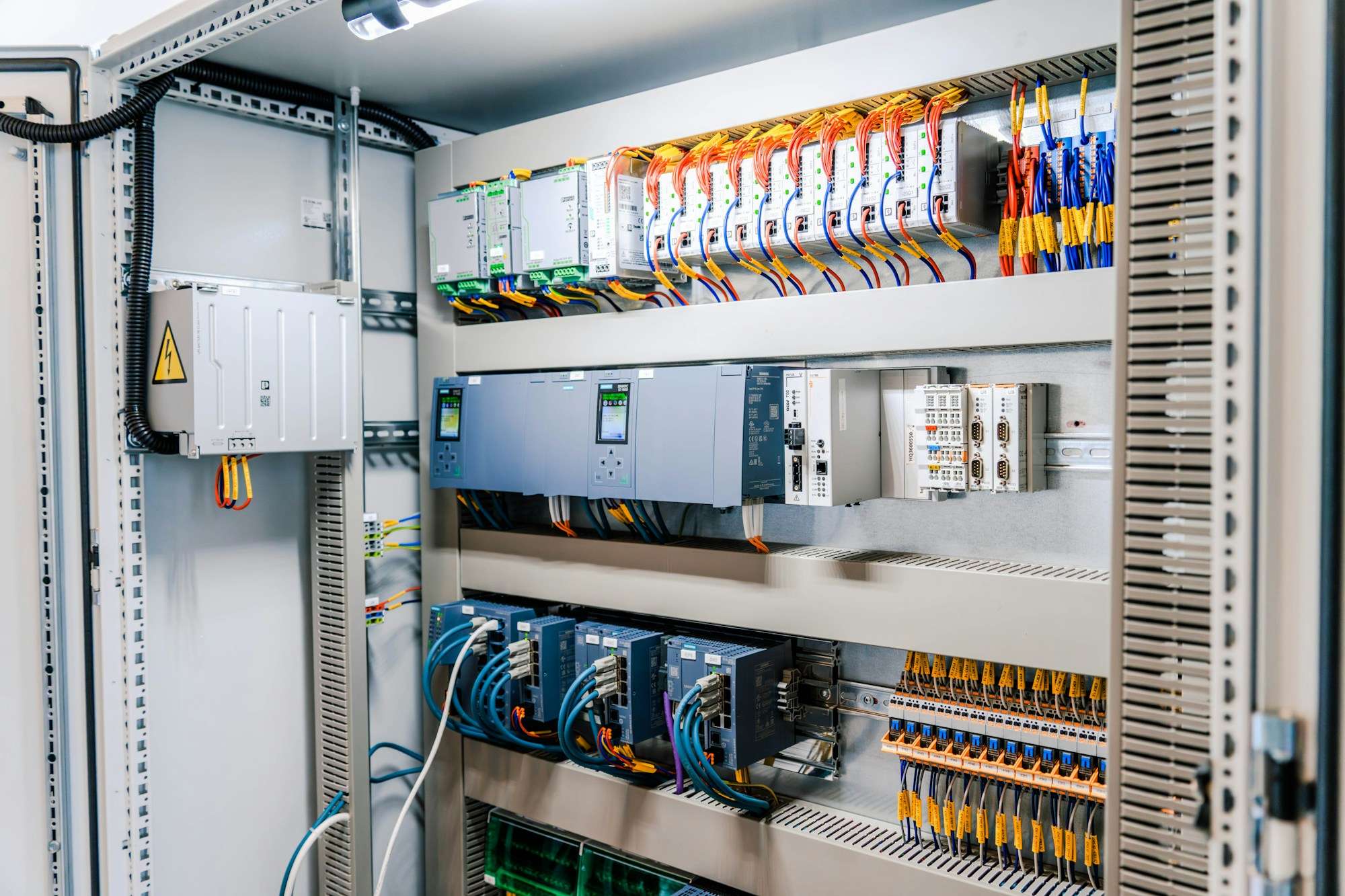 Open industrial control cabinet with PLC modules, network switches and neatly labelled wiring on DIN rails
